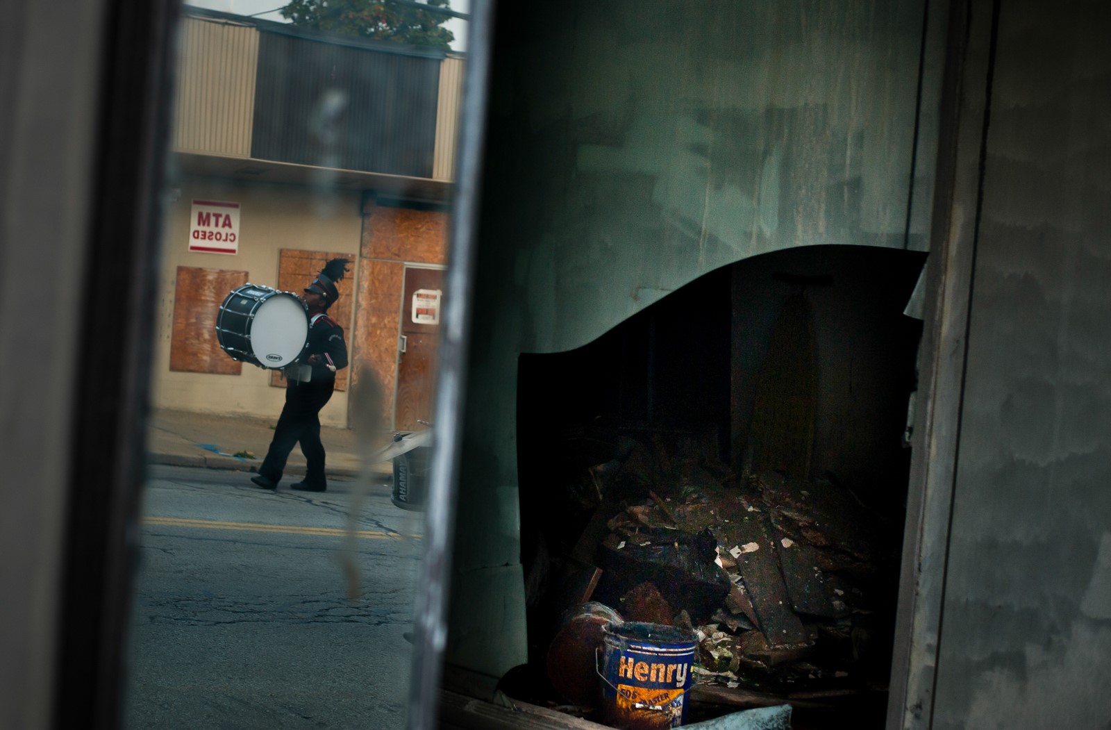 In the reflection of a shattered storefront, a marching band drummer walks down the street.