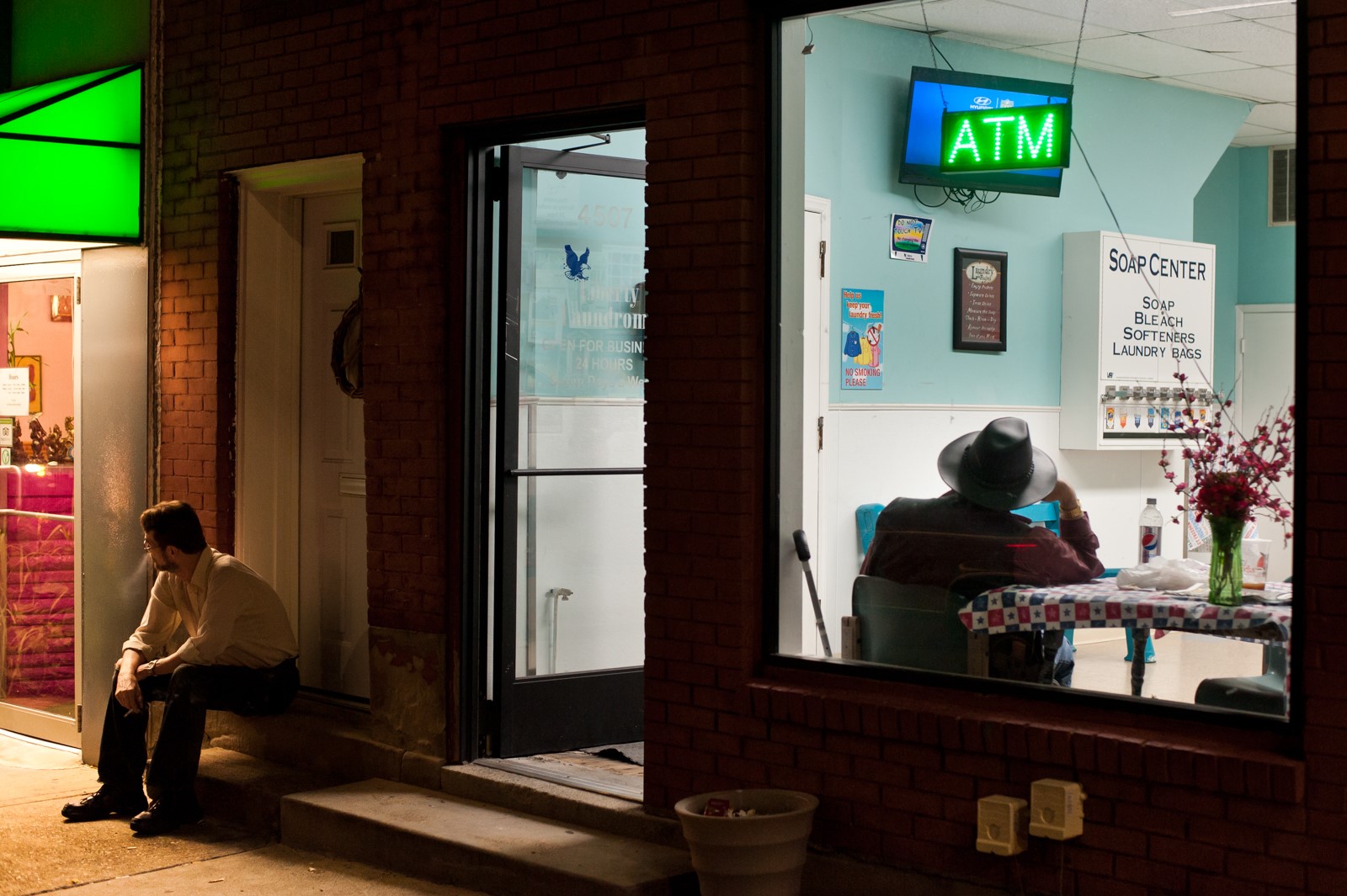 A man, smoking, sits on a stoop at night outside a well-lit laundromat.