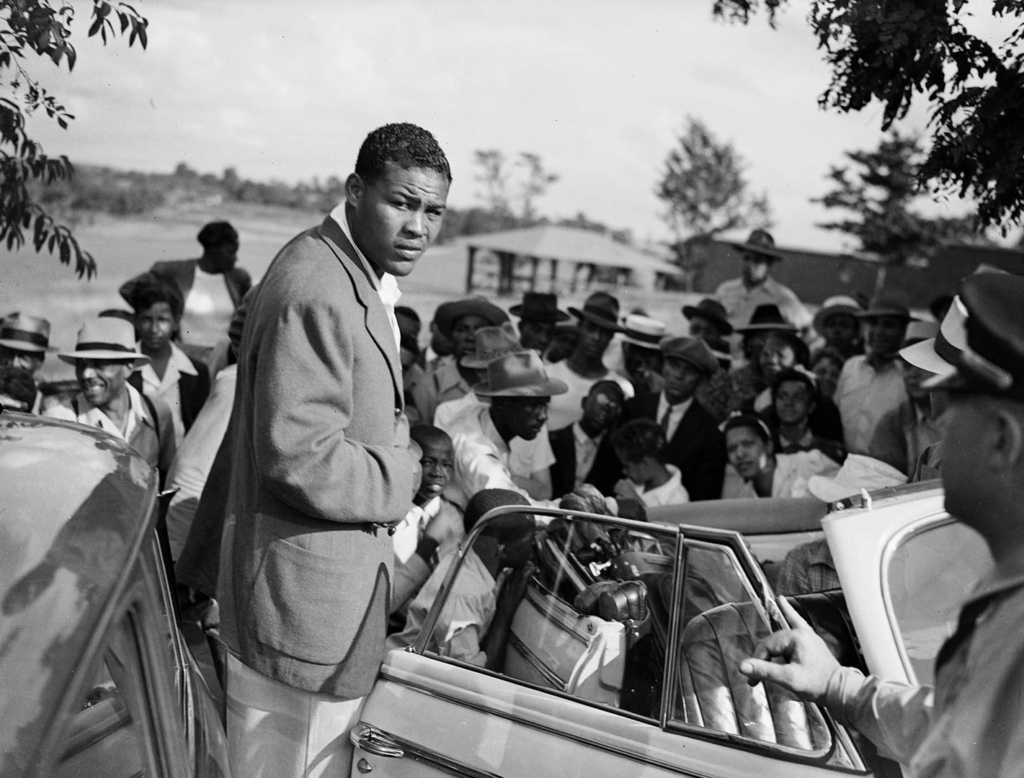 A well-dressed man stands next to a convertable; people crowd around in the background.