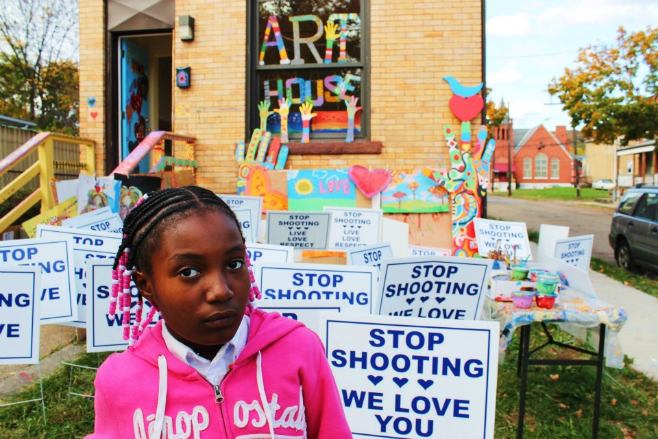 Girl in front of the art house with signs all over that say: Stop shooting we love you