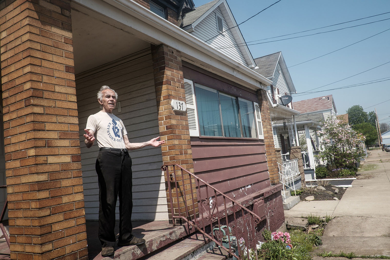 An elderly man stands outside his home, arms outstretched as he talks to the camera.