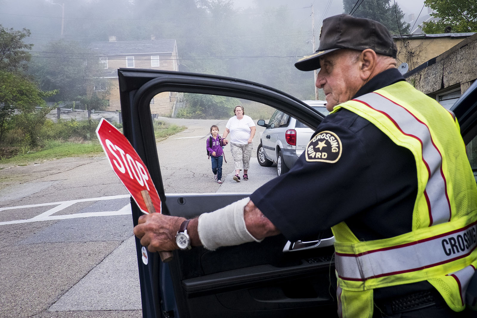 A crossing guard keeps watch; in the background, a mother walks her child to school.