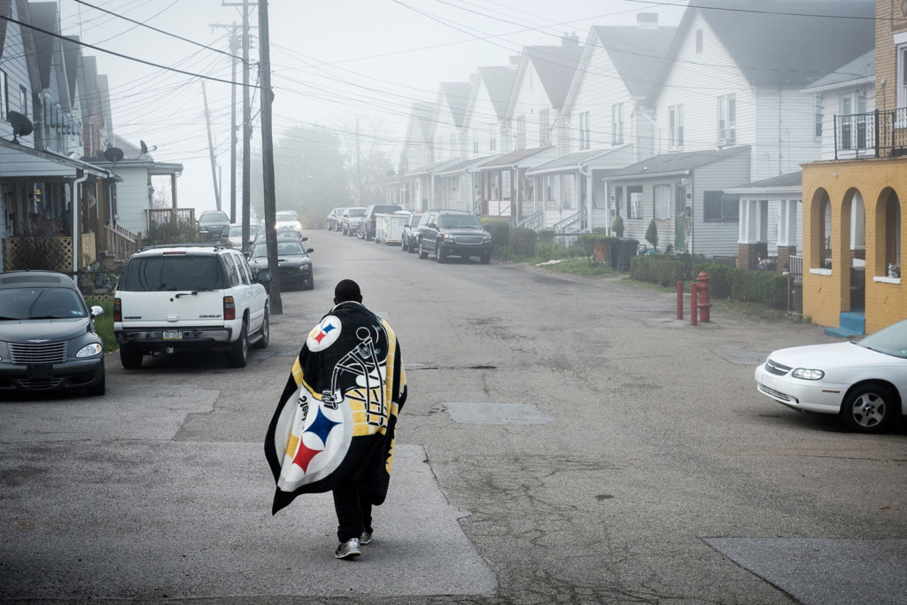 A man walks down a poorly-paved road, wearing a large Steelers blanket as a cape.