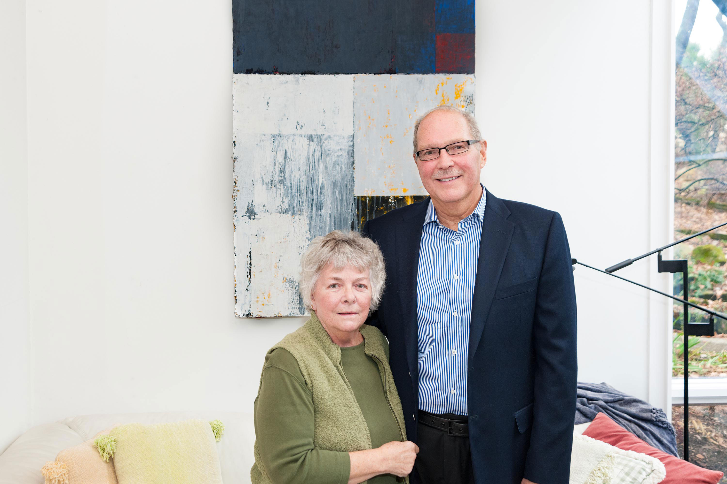 A man and woman, in their home, stand in front of a painting they have collected.