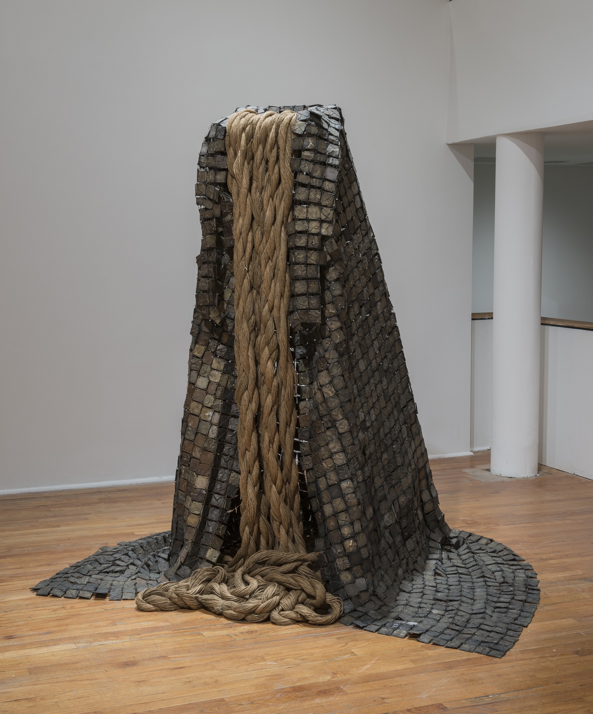 A tall sculpture made of black squares assembled together like scales, sweeping upward like a peak, with lengths of thick rope spilling over the edge like a waterfall