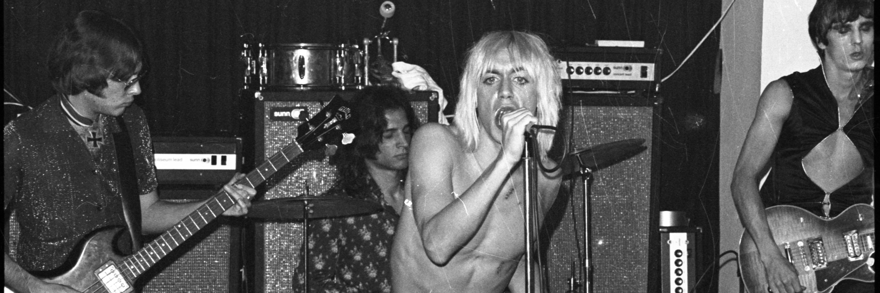 Rock and Roll band The Stooges, with two members looking away playing guitars, and singer Iggy Pop singing into a microphone and staring into the camera