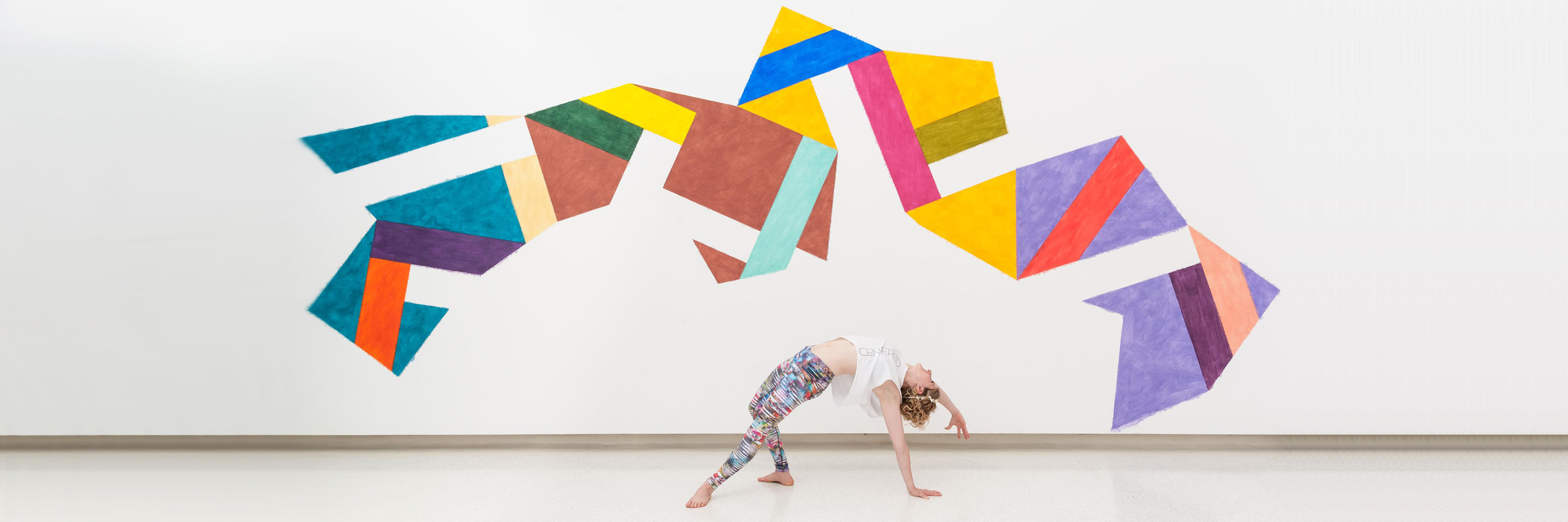 A woman doing a wheel pose in front of a colorful geometric artwork on a white wall.