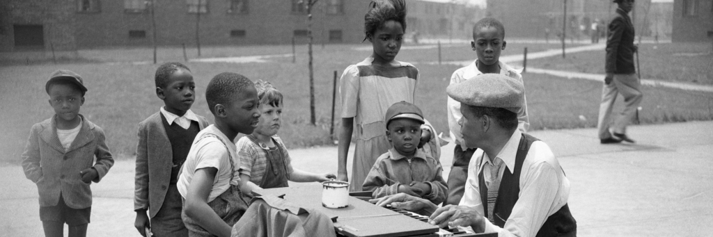 Children listening to man playing small piano on sidewalk in front of Terrace Village housing project in Pittsburgh’s Hill District