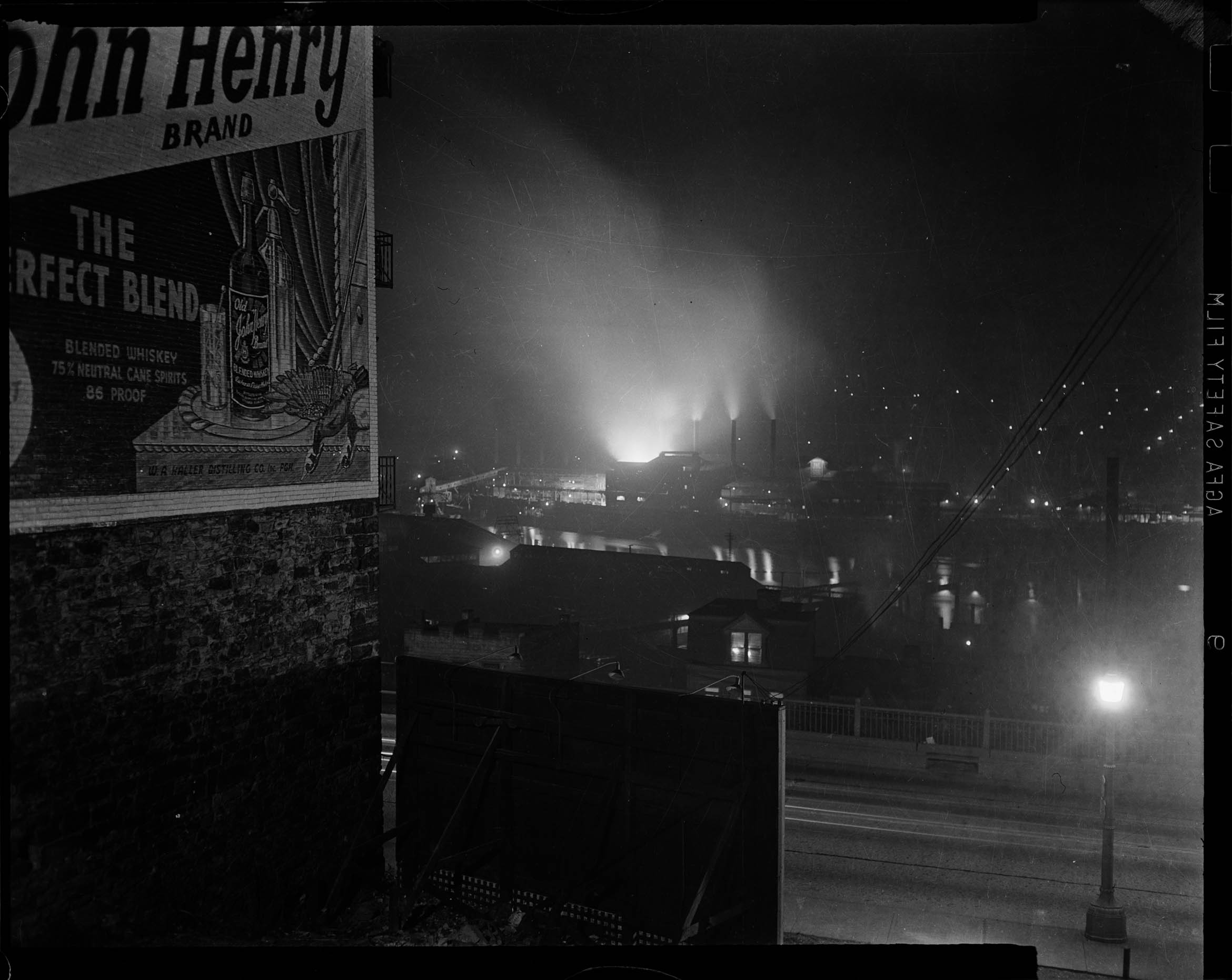 Night scene, with smokestacks of a factory in the background blasting out light and smoke. In the foreground on a building at left, a large wall mural shows a whiskey advertisement. It reads "John Henry Brand. The perfect blend."