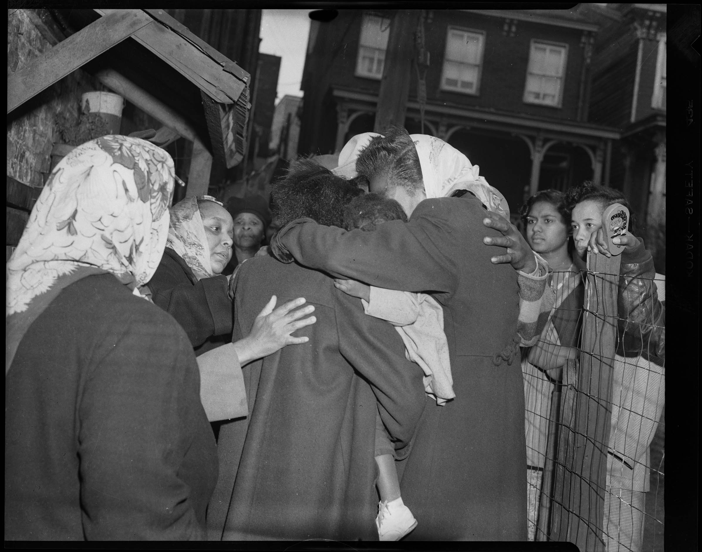 People embracing with buildings and several bystanders in background. The bystanders look concerned and upset.