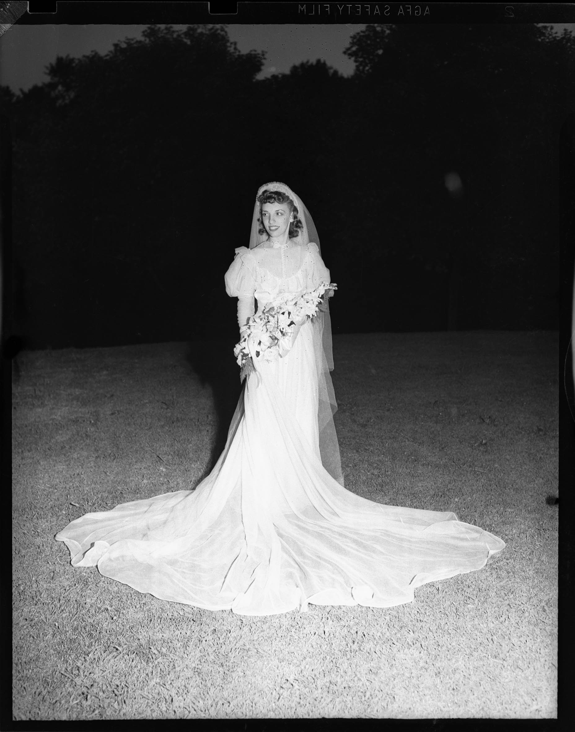 Portrait of a woman in a long wedding dress, which falls to the ground and fans out around her. She is holding a bouquet of flowers.