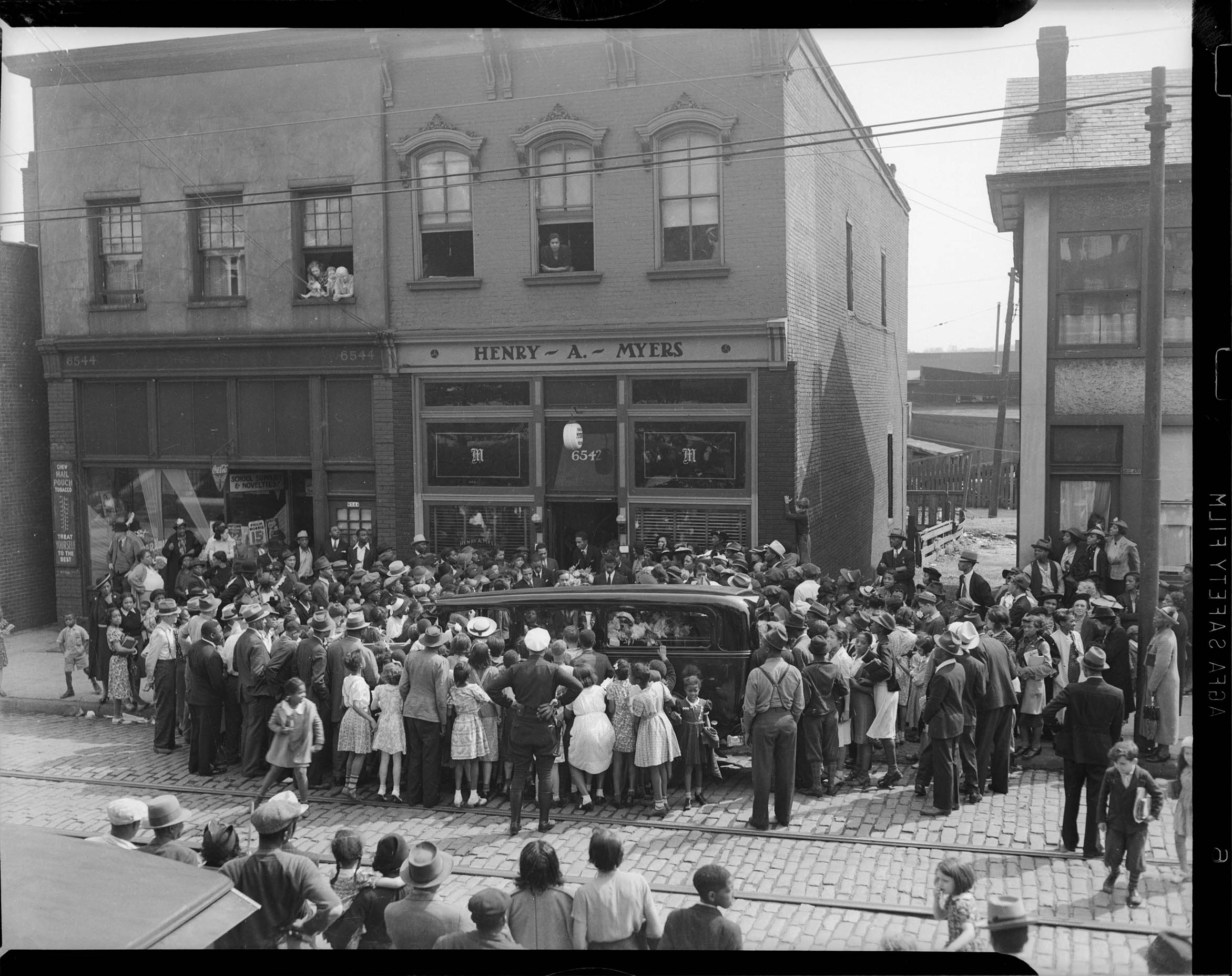 Busy city street, with large crowd gathered around a hearse