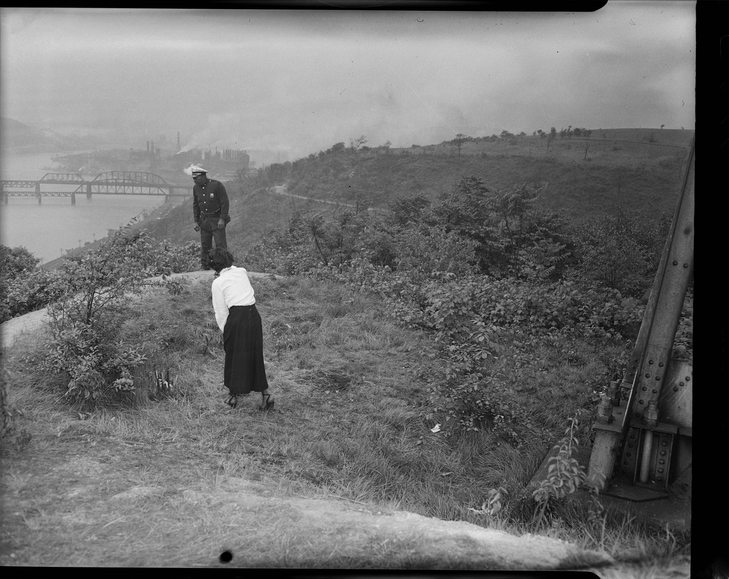 A police officer and a woman in a long skirt standing in a grassy area on top of a hill, looking over the edge of the hill