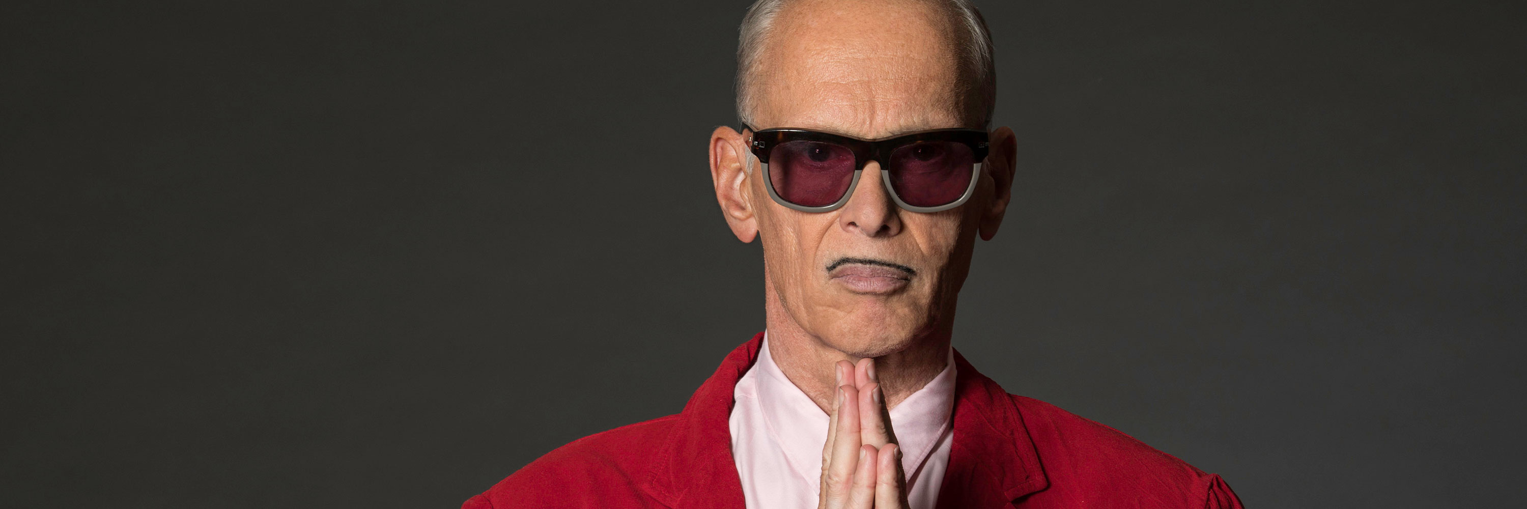 Photo of John Waters wearing a red suit and holding his hands together as if praying.
