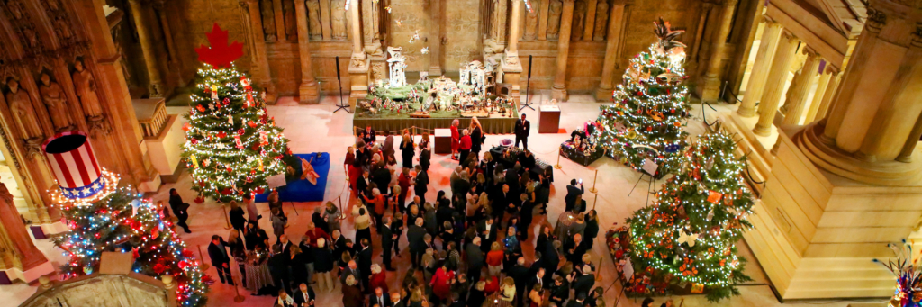 People celebrating in a grand museum hall among decorated holiday trees.