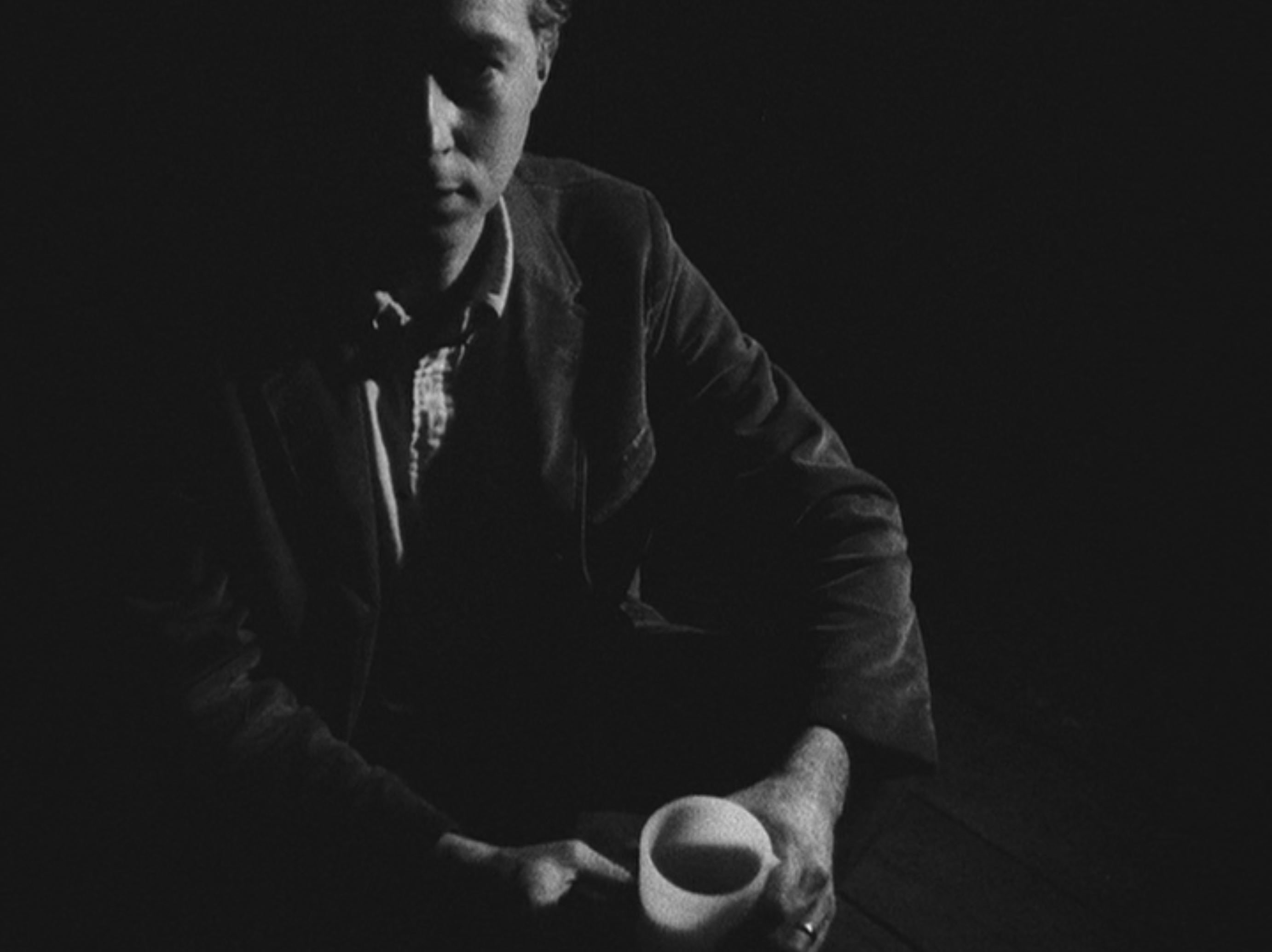 A black-and-white image of a man, apparently seated, holding a cup. Much of his face and body is shrouded in darkness.