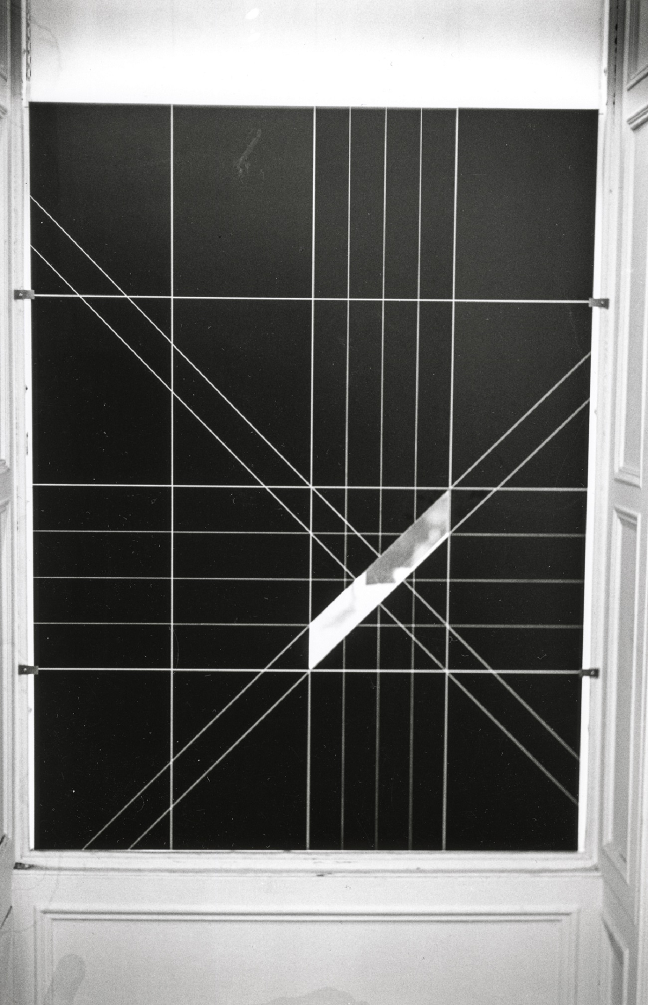 A dark-colored panel inscribed with light-colored gridlines and diagonals; this artwork has been placed over a window from the inside. A diagonal strip has been removed from the panel, admitting light from the outside.