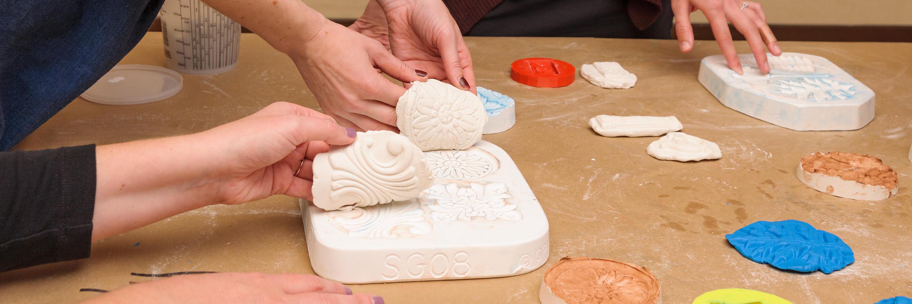Hands press clay into plaster impressions, copying the relief patterns
