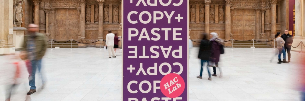 A long-exposure photo of a gallery space with blurry people walking by in the background. The focus is on a graphic reading Copy and Paste with a sticker reading HAC Lab