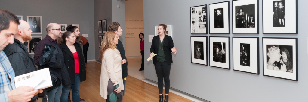 A woman stands next to photographs on a gallery wall, giving a tour to a group of visitors.