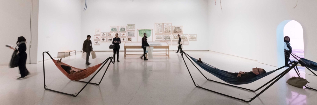 People walk around gallery and some lie down in hammocks.