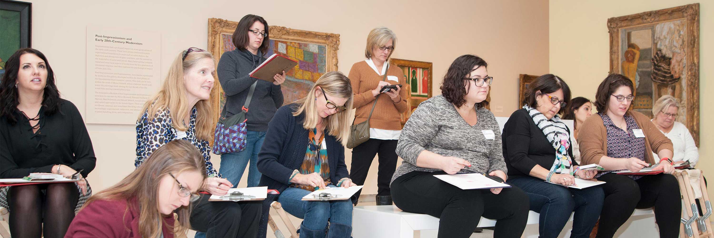 A group of women writing or drawing on clipboards inside of an art museum.