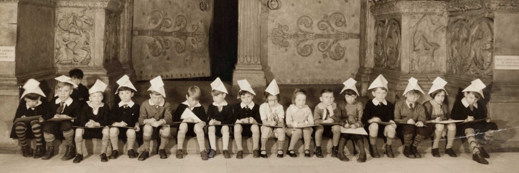 A group of young kids wearing school clothes and paper hats sit in a line in the Hall of Architecture.