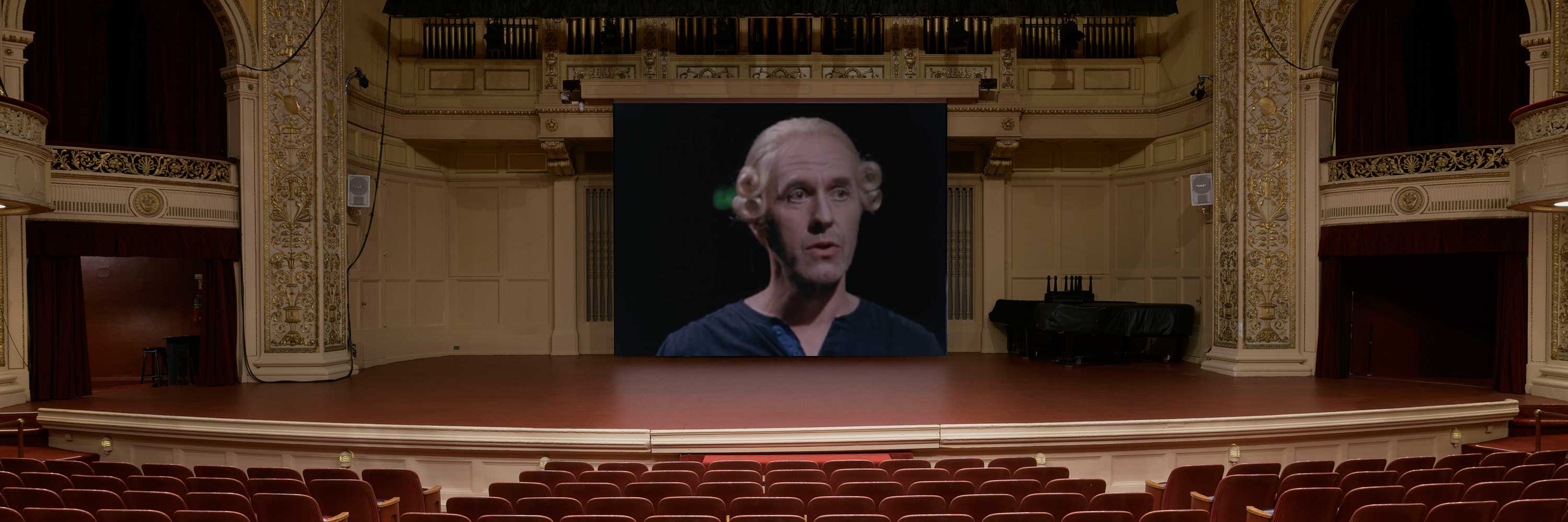 View of Carnegie Music Hall stage with projection screen of male with wig in conversation