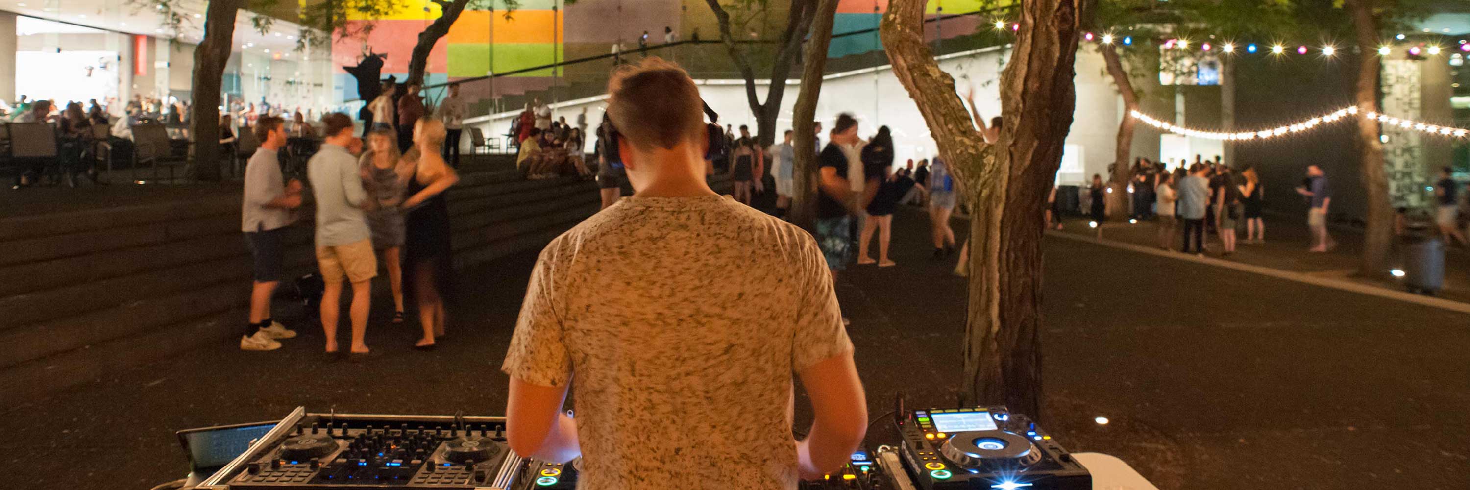 A man facing away DJ's for a crowd of people dancing outside.