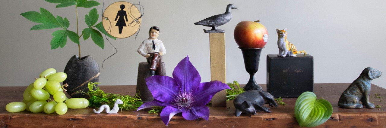 A table with carefully arranged objects: grapes, plants, a large purple flower, small figurines of people and animals, and an apple sitting in a cup.