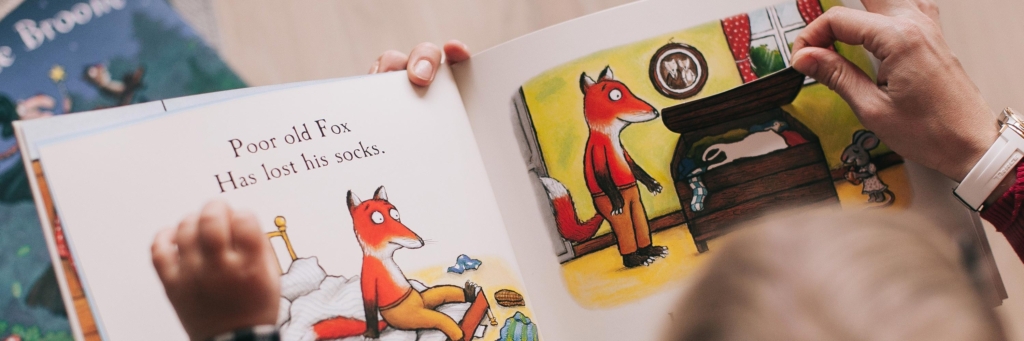A young child looks down at a picture book with a drawing of a fox, with the words "Poor old Fox has lost his socks"