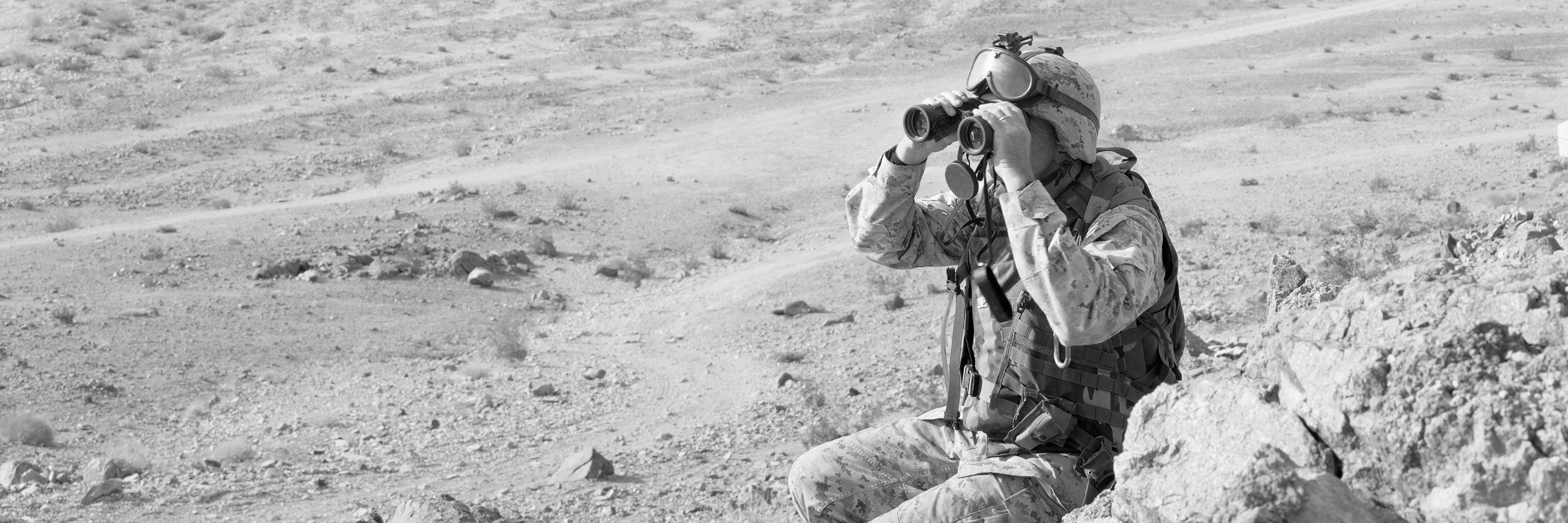 A black and white image of a men in tactical military gear holding binoculars, sitting in a desert landscape with dust and rocks