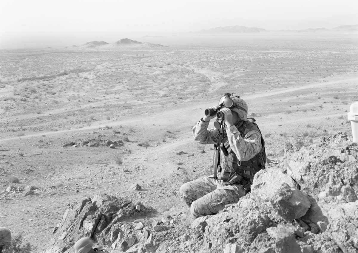 A man in military gear sits in a rocky landscape, looking through binoculars