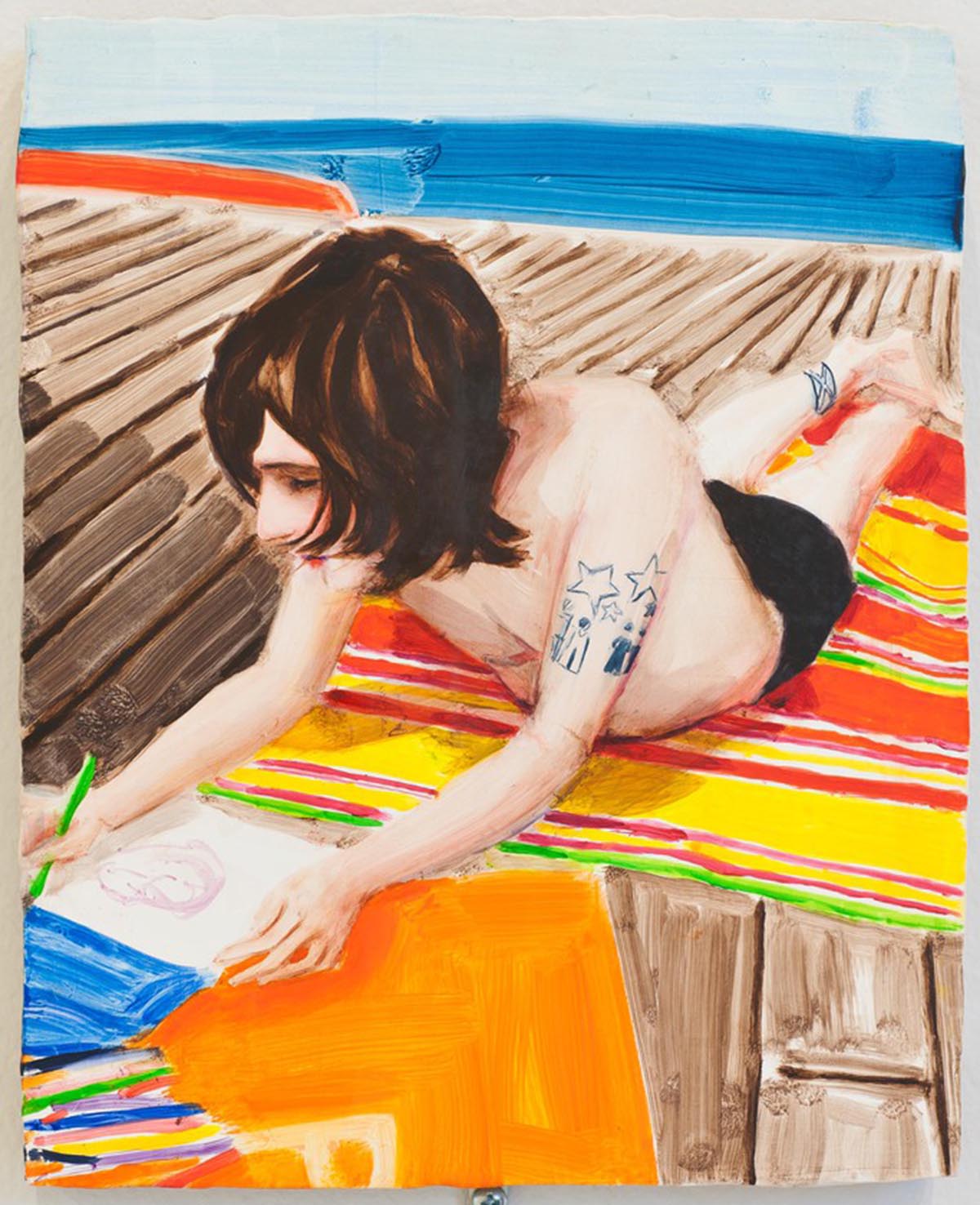 A young man wearing swim trunks lays on his stomach on a beach towel on a wooden boardwalk, drawing on a large piece of paper.