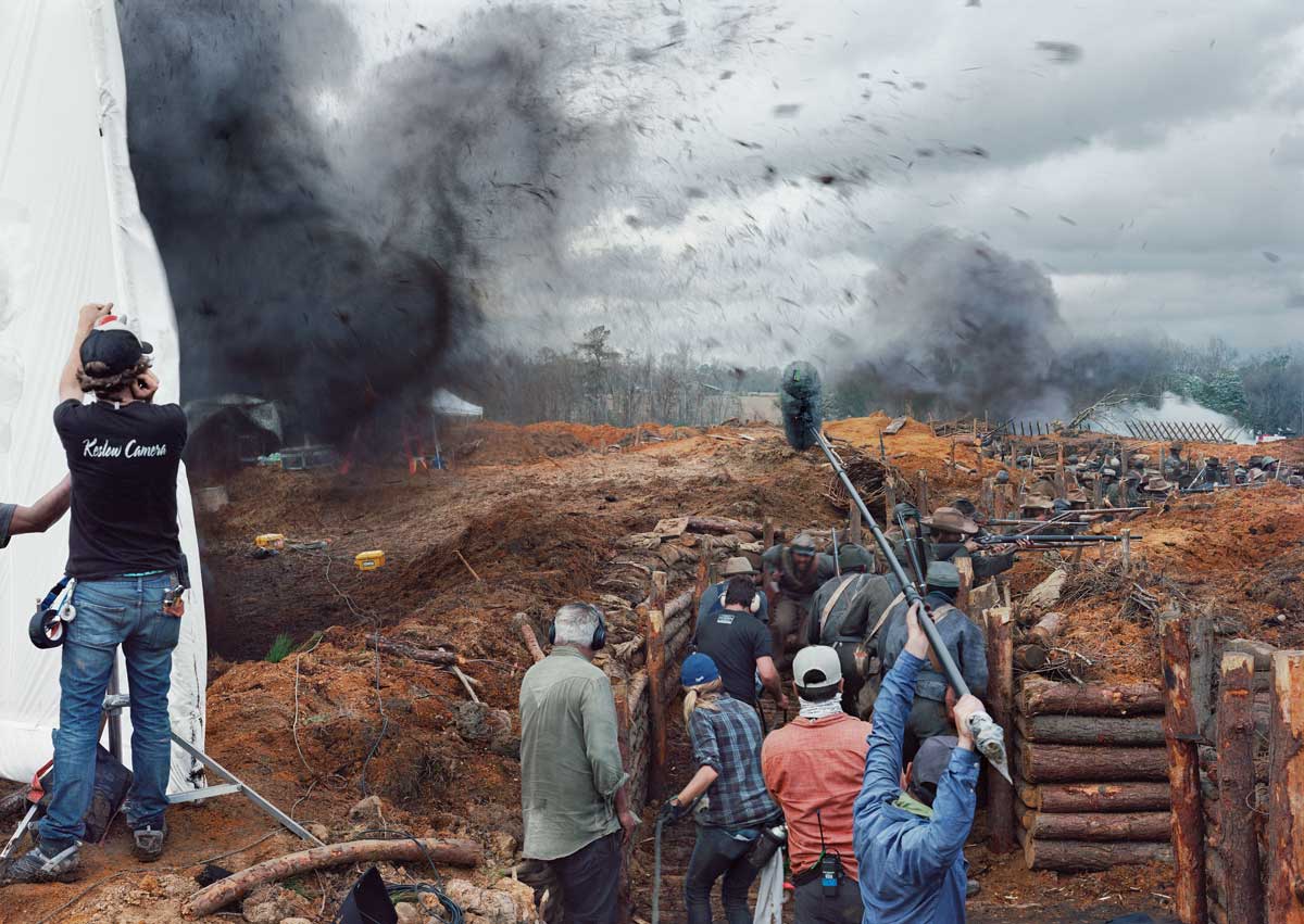 A group of people crouch in a trench, holding guns, in a dirty and chaotic environment. There are film crew members at the front of the image holding boom microphones