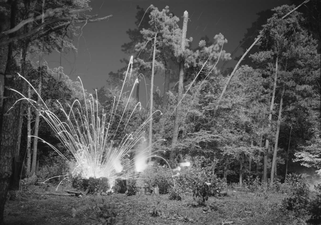 A black and white photo of tall trees and an explosion emanating from the ground