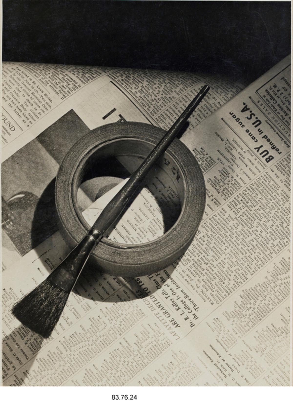 A black and white photo of a brush laid across a roll of tape, both sitting on top of a newspaper. 