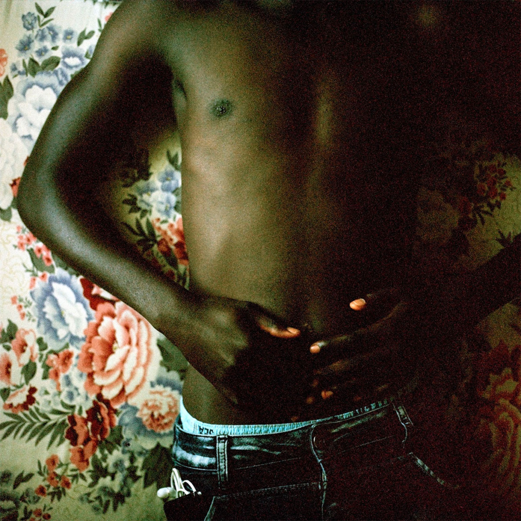 A close-up view of a person from shoulders to hips, relaxing against an old-fashioned floral backdrop wearing low-slung jeans and no shirt
