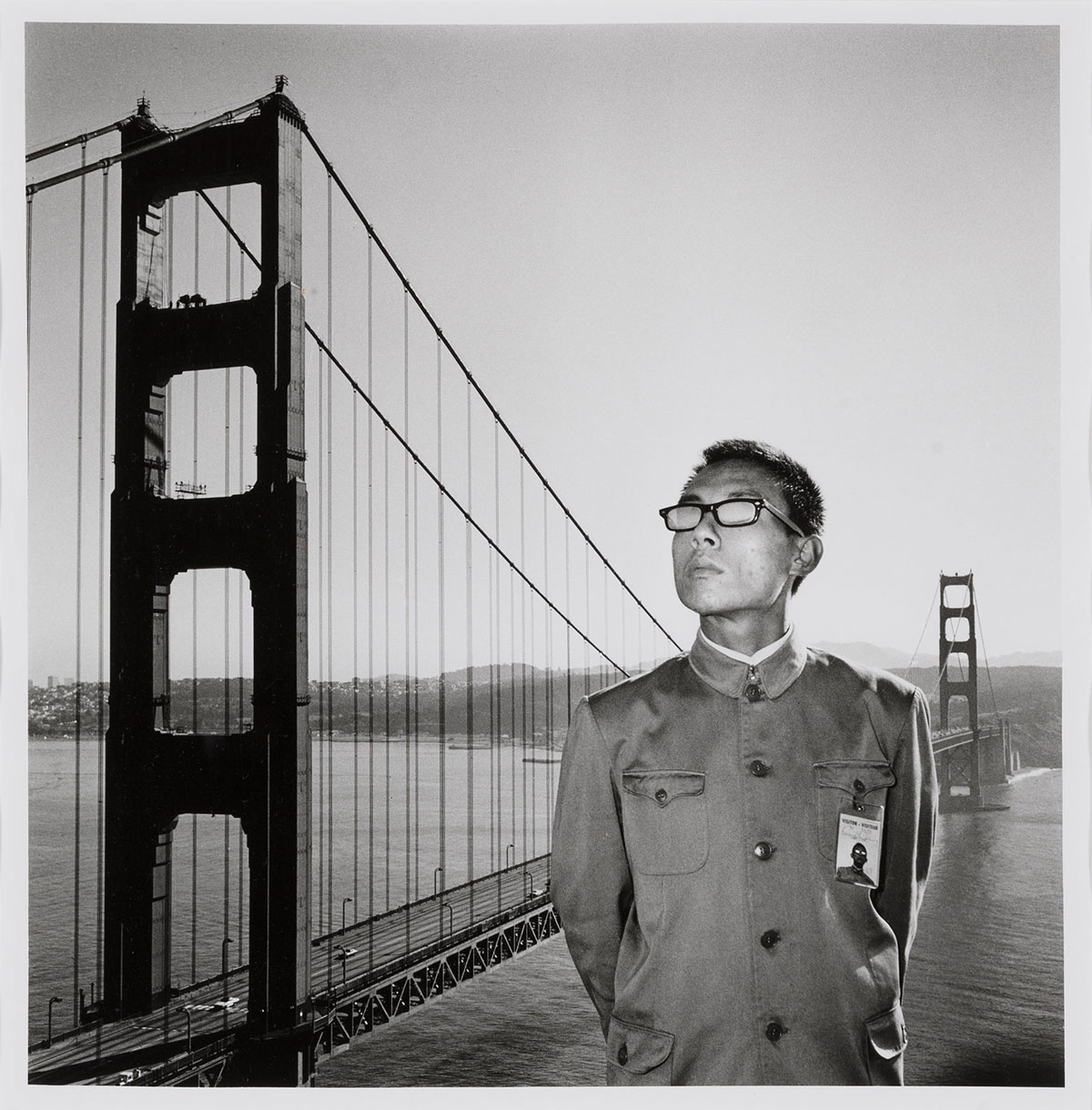 A man in a uniform-like jacket stands with his arms crossed behind him in front of the Golden Gate Bridge, looking out beyond the viewer,