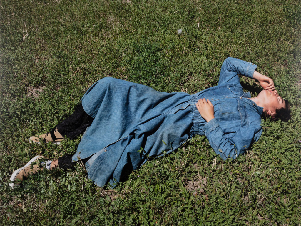 A color photograph depicting a person wearing a loose, long-sleeved denim dress stretches out on a grassy patch of ground