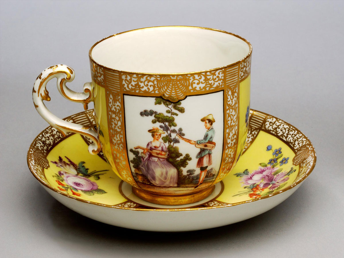 A gilded teacup and saucer with floral patterns. A panel showing a couple playing stringed instruments together appears on the side of the teacup