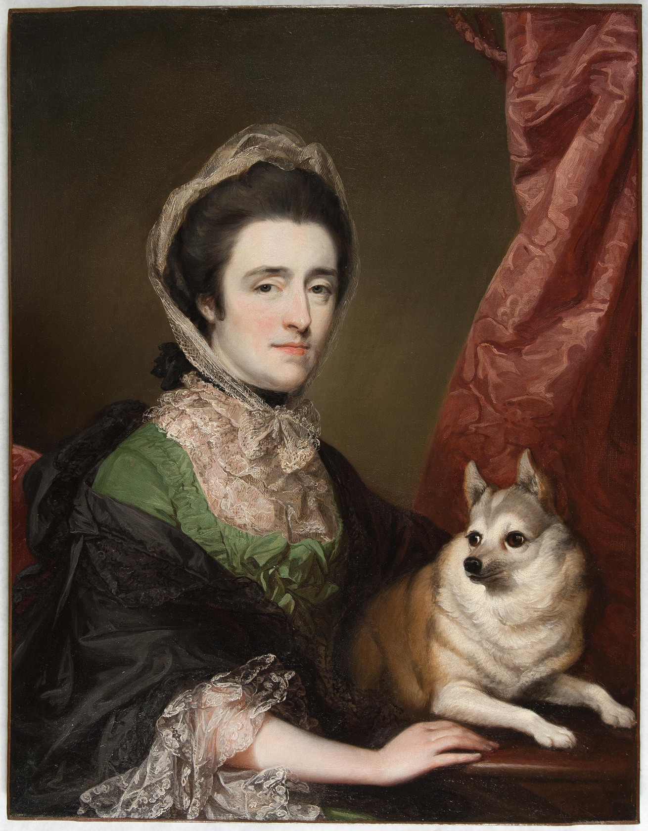 A woman wearing a shawl and hair covering sits with a small dog, looking at the viewer