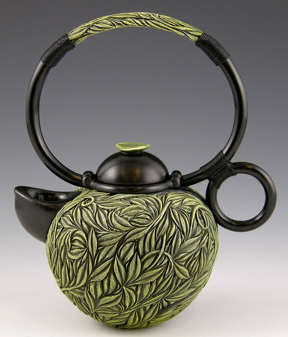 A teapot sculpture with a narrow, round handle attached to the cap; the body of the pot is etched with layers of green leaves