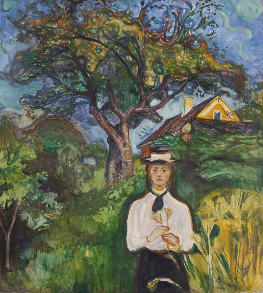 A young woman wearing a black and white dress and cap stands in front of a large tree with twisting branches. She is standing next to a bunch of wheat and holding some in her hands while looking directly at the viewer. The roofs of two houses can be seen in the background.