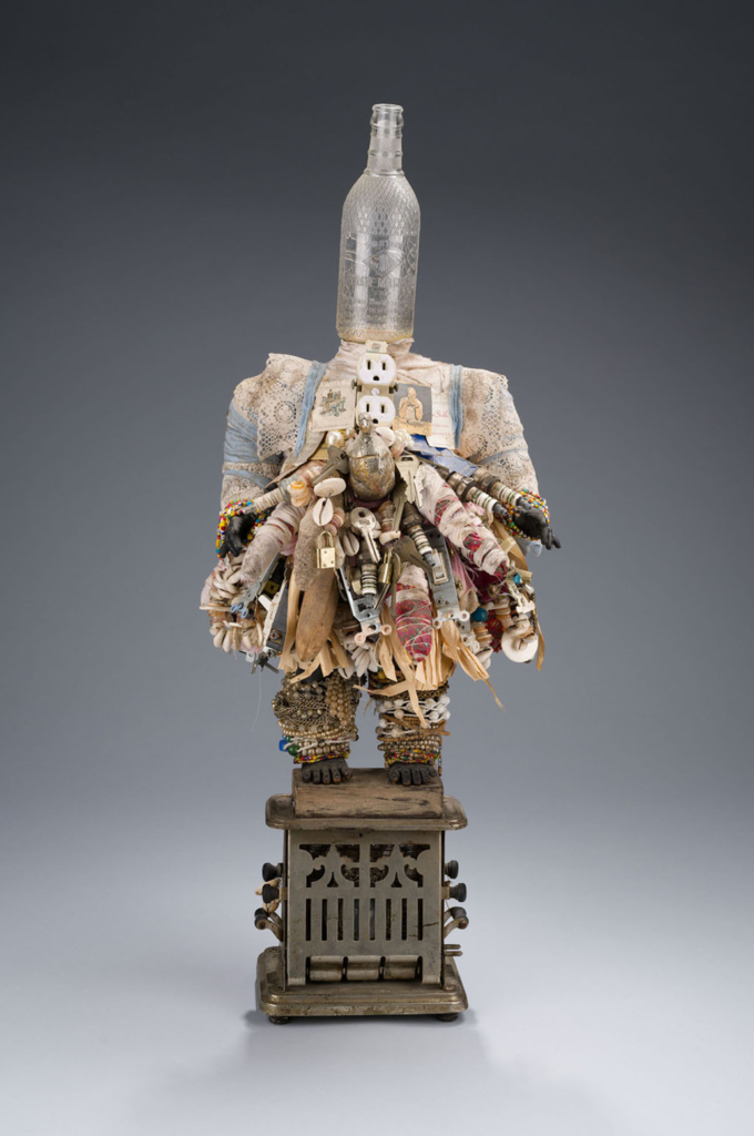 A sculpture depicts a human figure made from found objects, with a glass bottle forming the head, doll parts forming the limbs, and adornments made from items including beads, fabric, an electrical outlet, and the framed slave ownership photograph of a woman named Delilah.