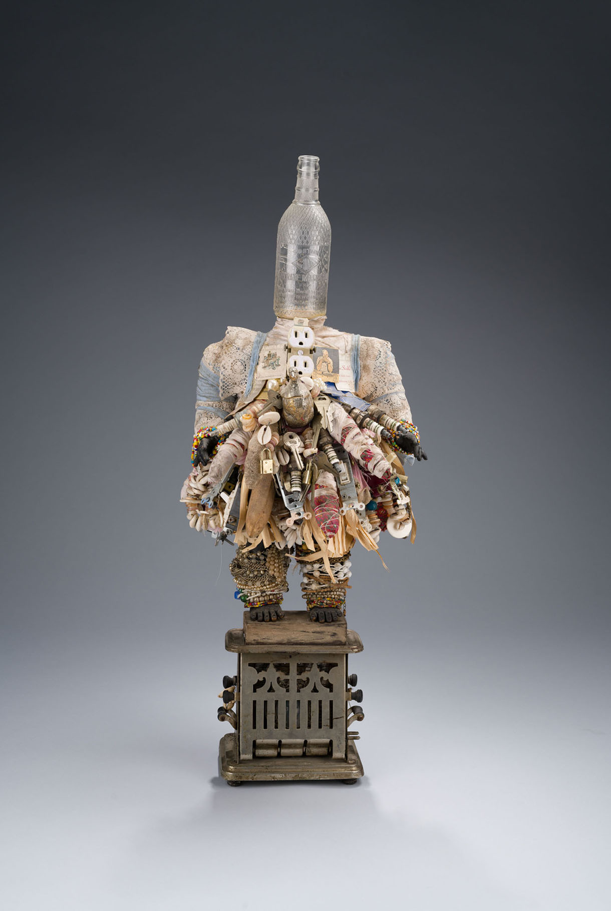 A sculpture depicts a human figure made from found objects, with a glass bottle forming the head, doll parts forming the limbs, and adornments made from items including beads, fabric, an electrical outlet, and the framed slave ownership photograph of a woman named Delilah.
