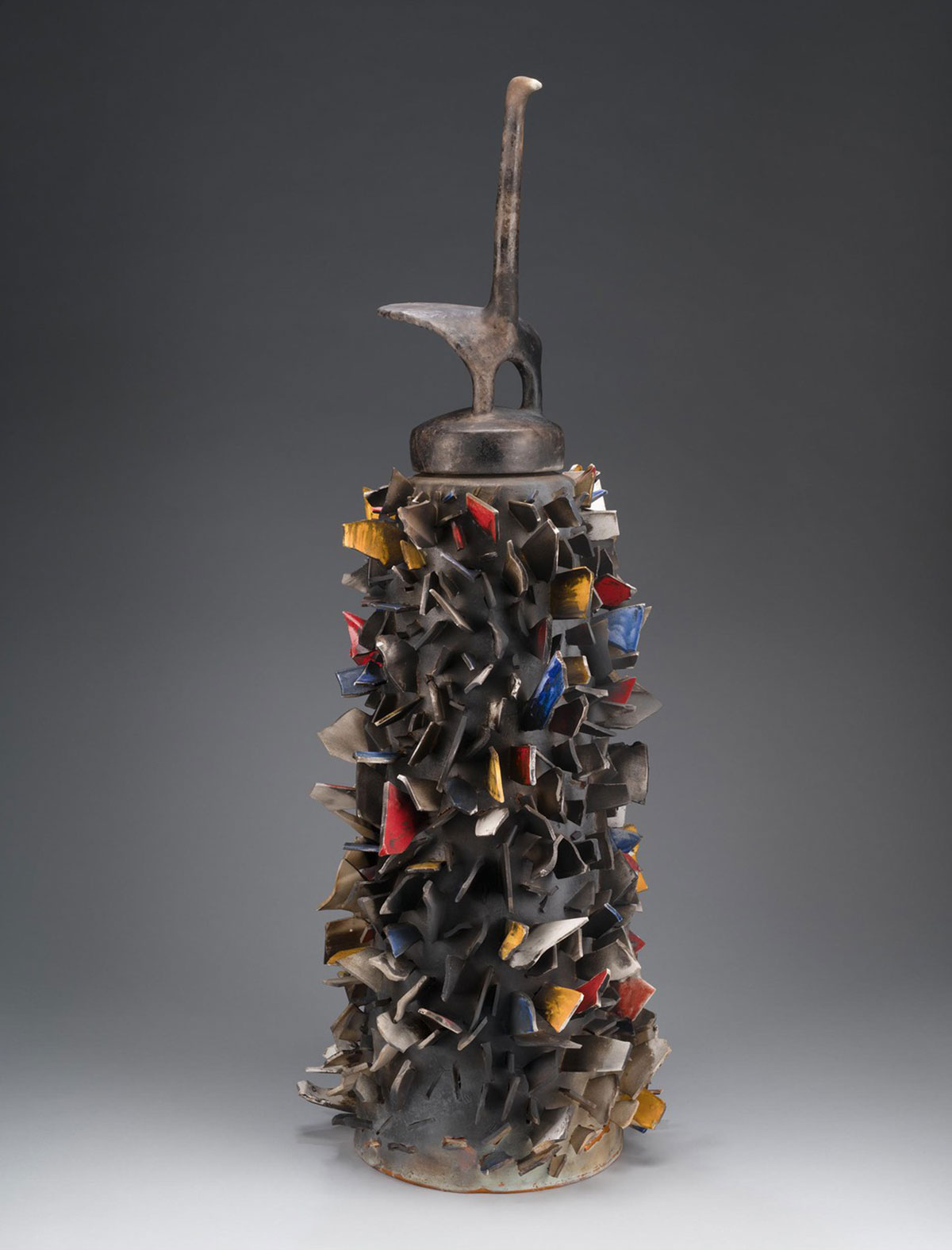 A tall sculpture with a narrow, cylindrical base covered in colored shards of porcelain; a tall earthenware figure sits atop the base