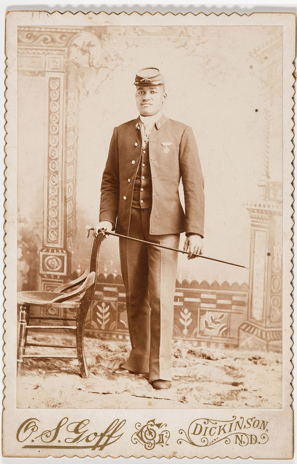 A man wearing an old-fashioned military uniform and hat stands in an ornate room next to a wooden chair, holding a walking cane across his body
