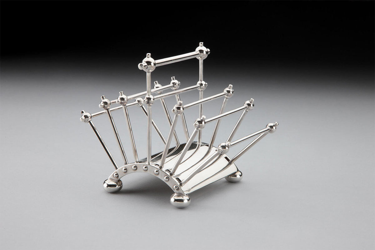 A silver kitchen device with a curved base supporting seven spindly arms that create six segments to hold a slice of toast