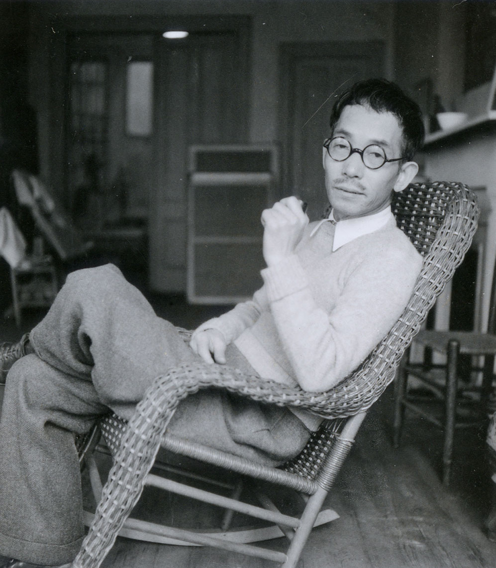 A young man wearing a sweater, collared shirt, and circular glasses leans back in a wicker chair inside a room